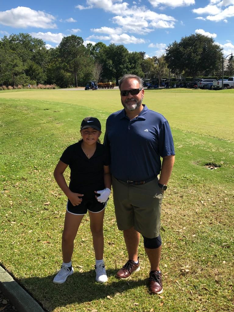 Father and daughter smiling together on a sunny golf course with the putting green behind them.