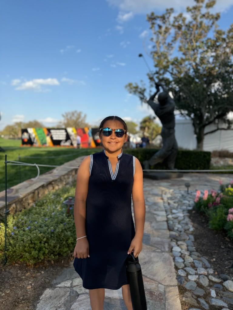 Young golfer in a navy dress posing in front of a golfer statue at an outdoor event.